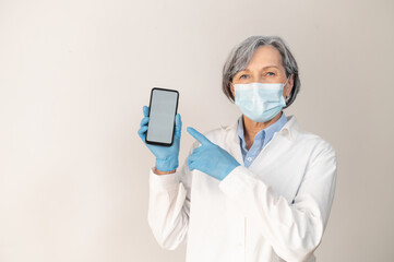 Fototapeta na wymiar Senior female doctor in a medical robe, protective face mask, and surgeon latex gloves posing isolated over gray background, pointing at the smartphone with a blank screen for advertisement