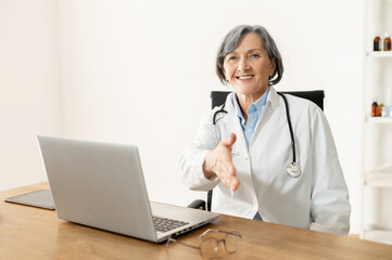 Senior old female doctor with stethoscope wearing lab coat sitting at the desk with laptop,...