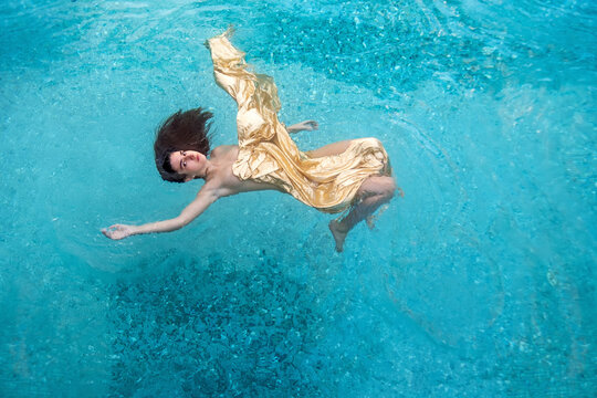 beautiful sexy young woman mermaid in gold dress, cloth floats weightless elegant in the blue turquoise spa pool water