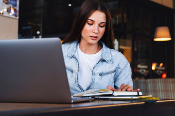 Fototapeta na wymiar Business woman sits at table in cafe in front of laptop, reads notes from notebook, makes notes in diary. Beautiful brunette female makes business calendar. Online training, remote work from home
