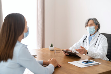 Senior mature female doctor in a medical robe and a face mask sitting at the desk, holding a touchpad, looking at the young lady patient, discussing medical results, digitized medical records concept