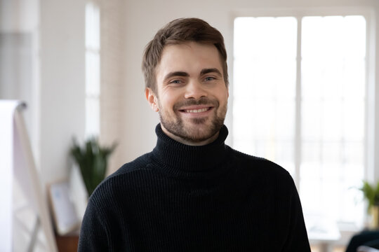 Head shot of happy male millennial posing in white interior. Profile picture of confident young man in black casual sweater looking at camera and smiling. Job candidate or employee corporate portrait