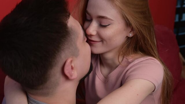 Close up video of a ginger couple kissing and embracing sitting on a red couch at home