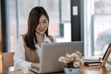 Young Beautiful smiling Asian business woman holding a coffee and laptop Placed at the wooden table at the office