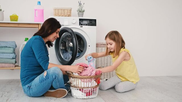 Mother and little daughter loading clothes for washing into machine sitting on floor in laundry room