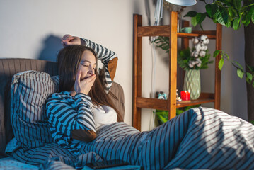 A young woman is lying in her cozy bed, stretching and yawning. Concept of bedtime pastime and rituals for a healthy and restful sleep