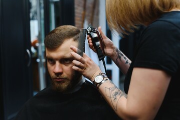 Professional hairdresser woman doing styling of handsome guy with electric hair clipper at barber shop