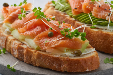 Two open sandwich, toast with salmon, cream cheese, avocado, cucumber slices on white concrete table. Morning healthy breakfast with fish and cup of drink, macro, close up