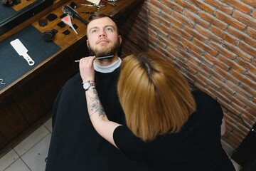 Skilled female barber cutting a beard of brunette adult guy in professional retro barbershop.