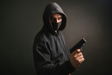 A gangster in a hood, a mask, holds a weapon in his hands on a dark background. The bandit aims...