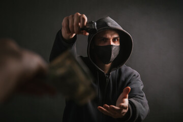 A gangster in a hood and a mask holds a weapon in his hands on a dark background and takes money from the victim. The bandit aims his pistol and demands dollars. Photo in the studio.