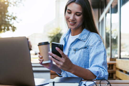 Young millennial woman working in cafe on open terrace, sitting in front of laptop, talking on the phone. Isolated woman learning distance learning, get knowledge over the internet. Online learning