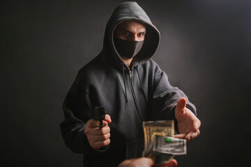 A gangster in a hood and a mask holds a weapon in his hands on a dark background and takes money...