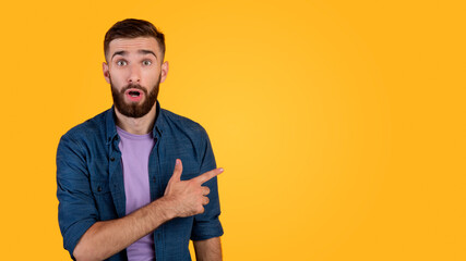 Shocked millennial guy pointing aside at empty space on orange studio background, panorama