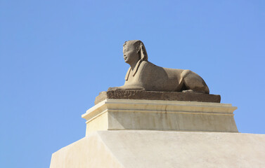 A sphinx in the northern Egyptian city of Alexandria on the railing of Pompey's Pillar. An ancient figure in the form of an animal with a human head. It is a clear day with blue skies.
