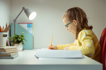 Pretteen schoolgirl in glasses studing at desk, writing and making homework at home during pandemic...