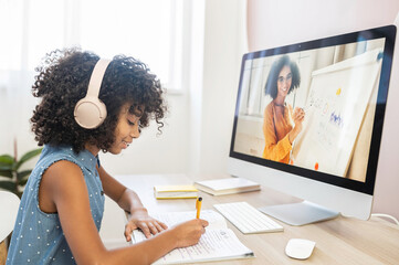 A clever multiethnic girl sits at table with a headphones, studying on the distance, doing homework, writes on workbook, watching online classes on the PC, side view. E-study, remote education - obrazy, fototapety, plakaty