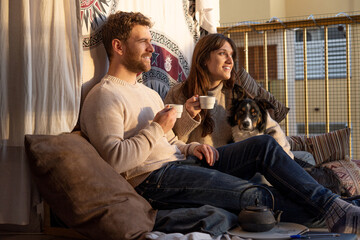 Young couple enjoying the sunset drinking tea on their cushion-filled balcony and their dog