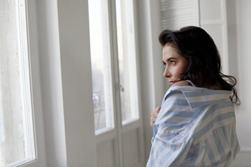 Side view of delicate female wearing domestic shirt standing in white room at home and looking away