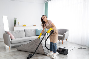 Positive female housekeeper using vacuum cleaner to tidy modern apartment. Sanitary service concept - obrazy, fototapety, plakaty