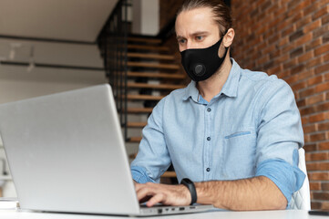 An male office employee wearing mask is using laptop at the loft style office. Social distance and safety at work during pandemic and viral disease