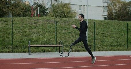 Side view of disabled male sportsman with amputated leg with prosthetic blade jogging at sports field. Handicapped male athlete at running track outdoors. Concept of health, sport, grid. - Powered by Adobe