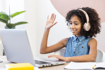 A pretty curly African schoolgirl wearing a headphones using a laptop for studying on the distance, a pretty biracial girl waving into webcam to classmates, to teacher or online tutor on the screen