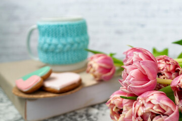 A white cup of tea in a knitted blue case stands on a book and pink flowers, tulips and gingerbread on a texture table. Spring concept.
