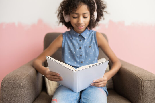 Smart biracial preteen school girl spending leisure time with a book reading sitting at armchair at home