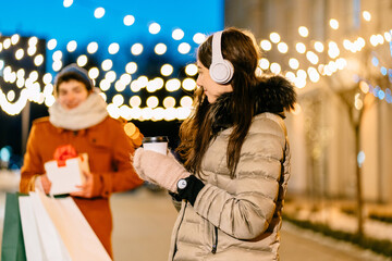 Fototapeta premium Happy young female woman in headphones warm clothes, paper bags, whitegift box with red bow waiting her boyfriend outdoor in winter city street. Winter holiday celebration concept.