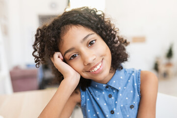 A close-up portrait of pretty African preteen girl, a schoolgirl wearing dot shirt looks at camera sitting at the desk at home