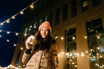 Joyful young female in warm outfit drinking warm coffee using sparkles for celebrating winter holidays. Two women having fun outdoors with garland light around in night city street.