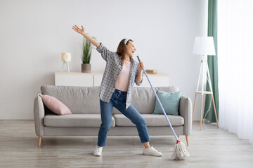 Happy woman singing while washing floor at living room, using mop as microphone, having fun during house chores