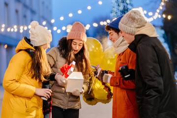 Group happy men and women standing outdoor with presents box, coffee cups and balloons in decorated...