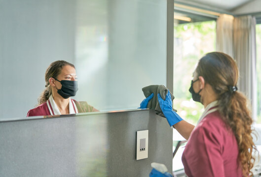 Female hotel maid in face mask and gloves cleaning bathroom mirror