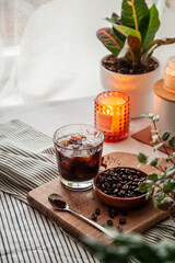 A glass of iced coffee on a wooden cutting board and a striped towel, coffee beans scattered around, on the background home staying plants and cozy candles