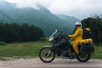 A girl in a yellow raincoat, shoe covers and a white helmet. Motorcyclism and travel. Sightseeing tour. Top of the Mountains. A gray day with thunderclouds. Copy space, biker's outfit.