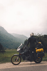 Fototapeta na wymiar A motorcycle driver with motorbike, Adventure vacation, biker dressed in raincoat. sealed bag, water resistant, overalls. Mountains road trip, side bags equipment. copy space. Vertical photo