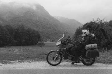 Obraz na płótnie Canvas A motorcycle driver with motorbike take a rest, Adventure vacation, biker dressed in raincoat. sealed bag, water resistant, overalls. Mountains road trip, side bags equipment. black and white