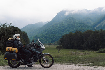 Fototapeta na wymiar A motorcycle driver with motorbike take a rest, Adventure vacation, biker dressed in raincoat. sealed bag, water resistant, overalls. Mountains road trip, side bags equipment. copy space