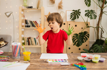 surprised little boy artist draws with his hands on white paper at a table in a room