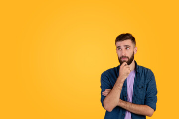 Portrait of attractive young guy thinking and touching his chin over orange studio background, copy...