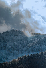 The clouds break up over a snow-covered, forested hills.