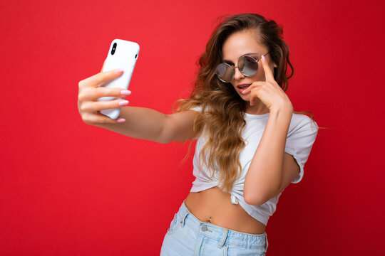 amazing beautiful young blonde woman holding mobile phone taking selfie photo using smartphone camera wearing sunglasses everyday stylish outfit isolated over colorful wall background looking at