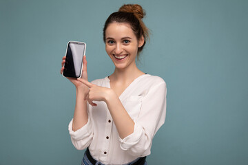 Naklejka premium Photo of beautiful smiling young woman good looking wearing casual stylish outfit standing isolated on background with copy space holding smartphone showing phone in hand with empty screen display for