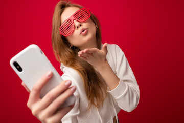 Naklejka premium Closeup portrait of attractive positive smiling young blonde woman wearing stylish white hoodie and funny colorful glasses standing isolated over colourful background holding and using mobile phone