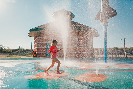 Cute adorable Caucasian funny girl playing on splash pad playground on summer day. Happy child having fun in water. Seasonal water sport recreational activity for kids outdoors.