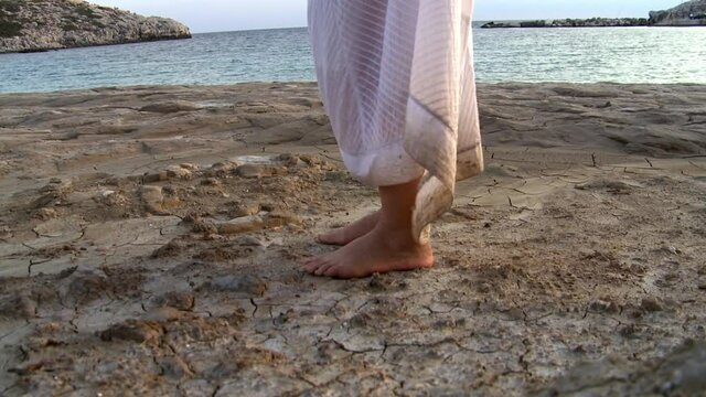 A woman alone in a white dress on a deserted beach walks on a mud path and dry land next to the sea 