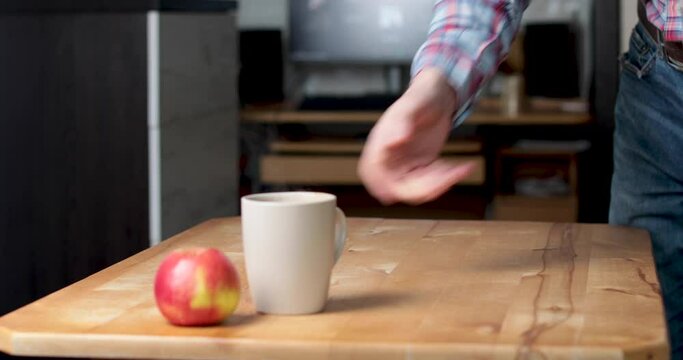Office worker in formal wear jeans and checkered shirt enter the room with laptop and sitting on his workplace with wooden table and cup of coffee and apple on it. 