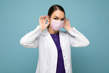 Photo of beautiful young woman doctor in white coat and medical mask standing isolated over blue background. Coronavirus concept